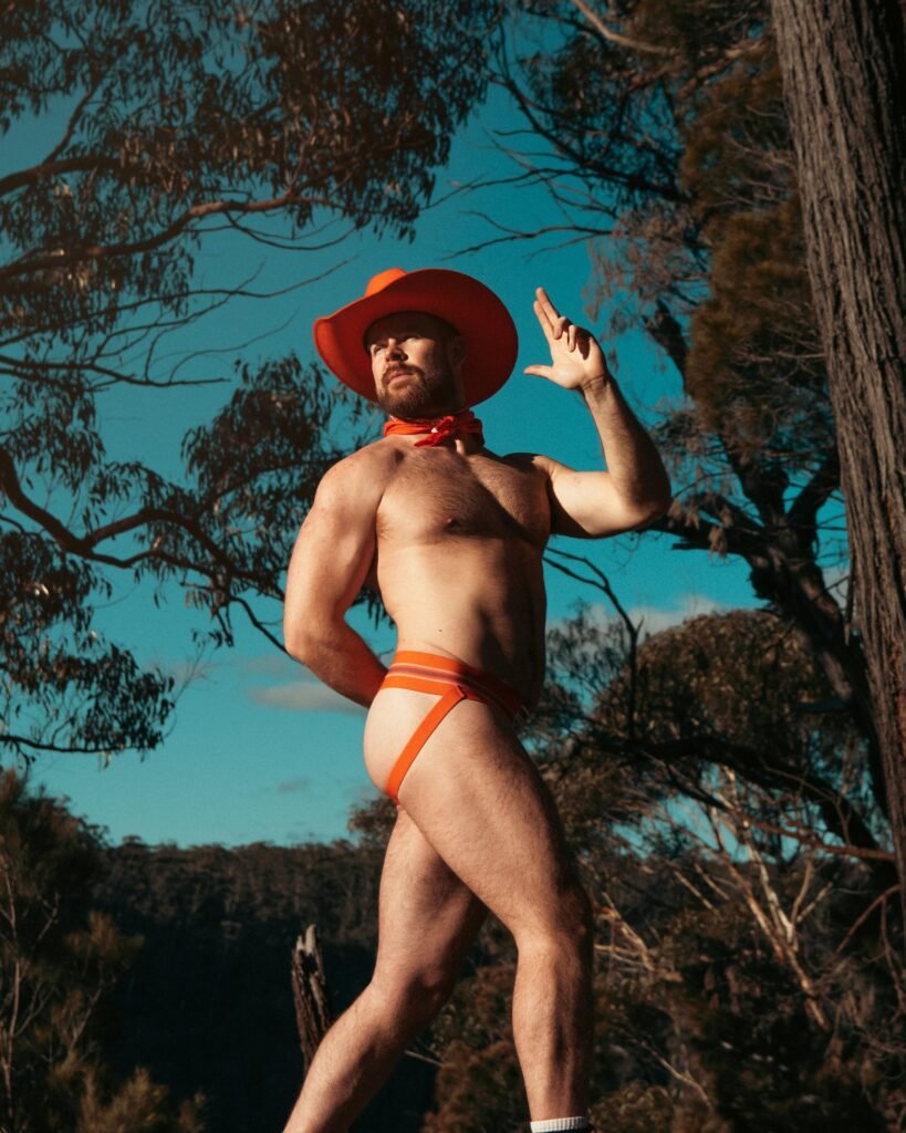 Gabe Woods in an orange cowboy hat and jockstrap standing in Australian bushland under a blue sky