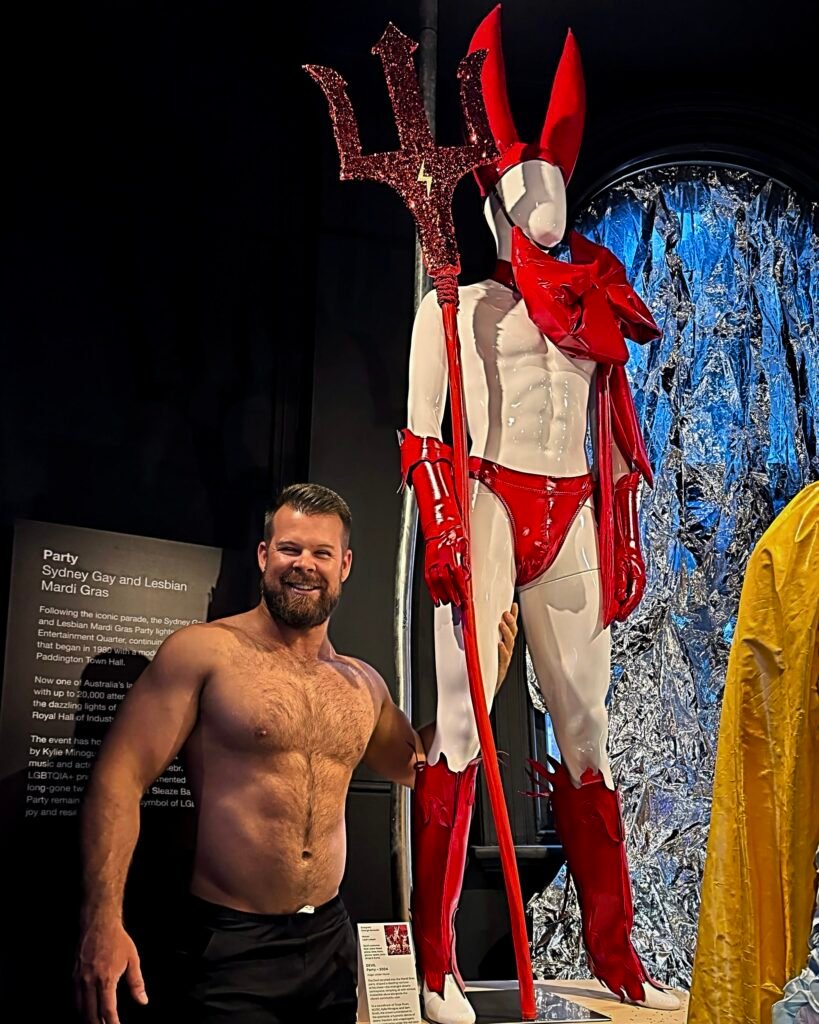 Gabe Woods, shirtless and smiling, beside a mannequin in the red Devil costume from the 2024 Mardi Gras Party display in The Making of Mardi Gras exhibition at Qtopia Sydney