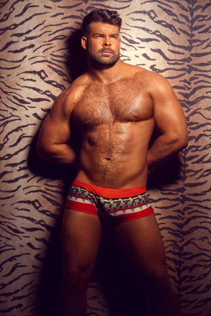 Gabe Woods standing against a tiger-print wall in red geometric-print swim trunks, warm studio lighting, photographed by Christian Scott