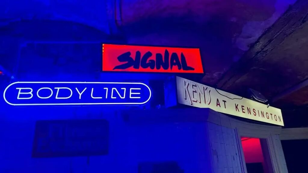Neon and lightbox signs for Signal, Bodyline and Ken's at Kensington glowing under blue light in The Underground exhibition at Qtopia Sydney